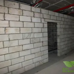 Wnętrze w trakcie budowy: ściana z białych bloczków, surowy strop z instalacjami, otwór drzwiowy, logo firmy w rogu.