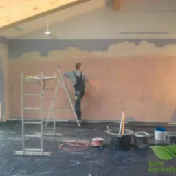 Pracownik w kombinezonie na drabinie wygładza szpachelką nierówną, pomarańczowo-beżową ścianę w pomieszczeniu z zabezpieczoną folią podłogą. Widoczne drabiny, wiadra z narzędziami i logo firmy Bami...