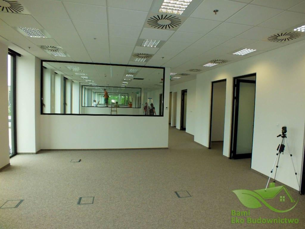 Wnętrze biurowe z wykładziną dywanową, białymi ścianami, sufitem podwieszanym i dużym lustrem, w którym odbijają się osoby pracujące na drabinie, w rogu stoi statyw z kamerą, widoczne logo firmy...