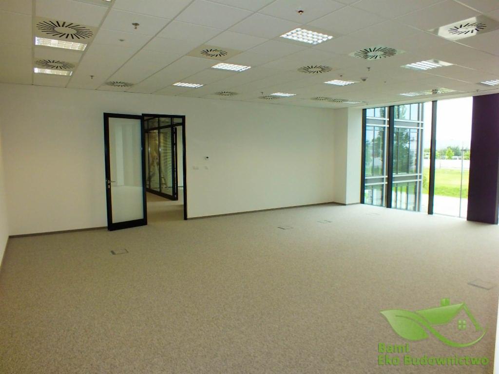 Przestronne, jasne wnętrze biurowe z beżową wykładziną, przeszklonymi ścianami i otwartymi drzwiami do kolejnych pomieszczeń, widok na zieleń za oknami, sufit kasetonowy z równomiernie...