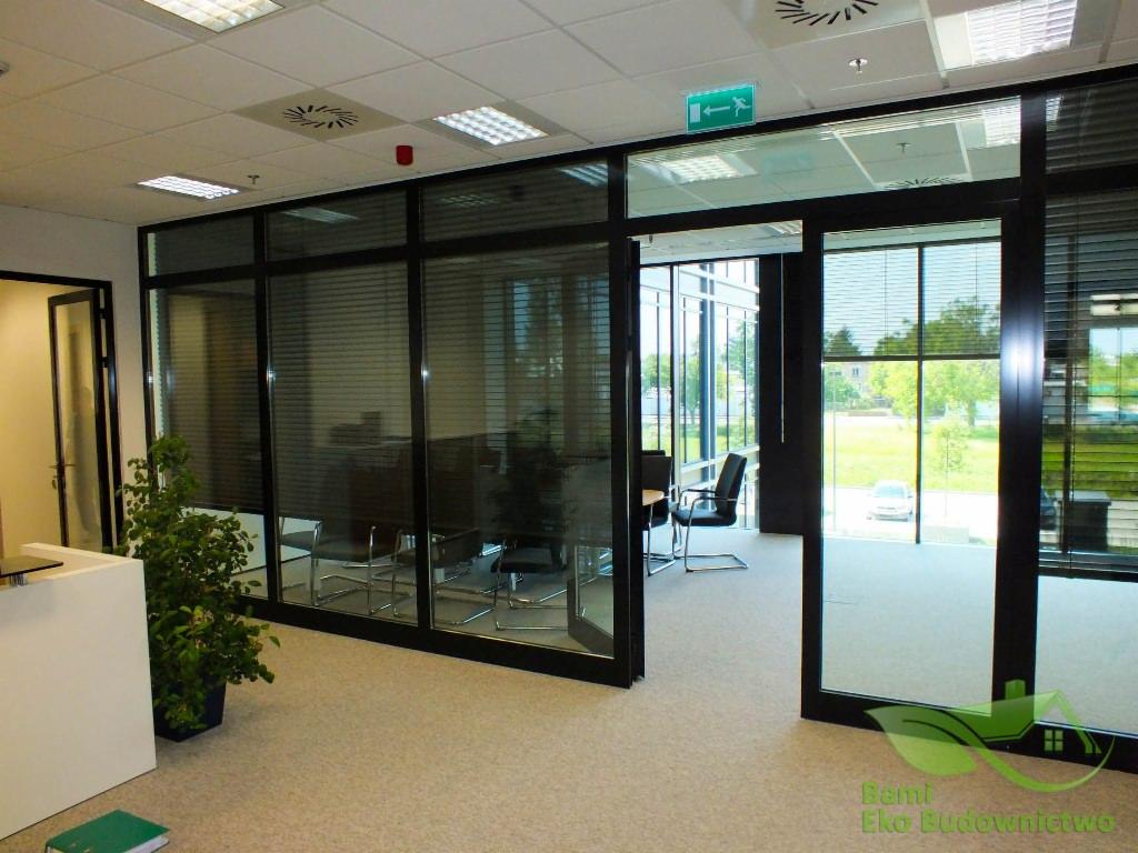 Nowoczesne biuro z przeszklonymi ścianami działowymi i roletami, widok na zewnątrz, minimalistyczny wystrój, jasna wykładzina, roślina doniczkowa przy recepcji, logo firmy w prawym dolnym rogu.