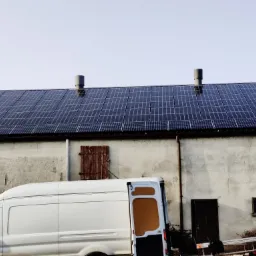 Instalacja paneli słonecznych na dachu budynku gospodarczego w Zagórowie, widoczny biały samochód dostawczy i drabina oparta o ścianę.