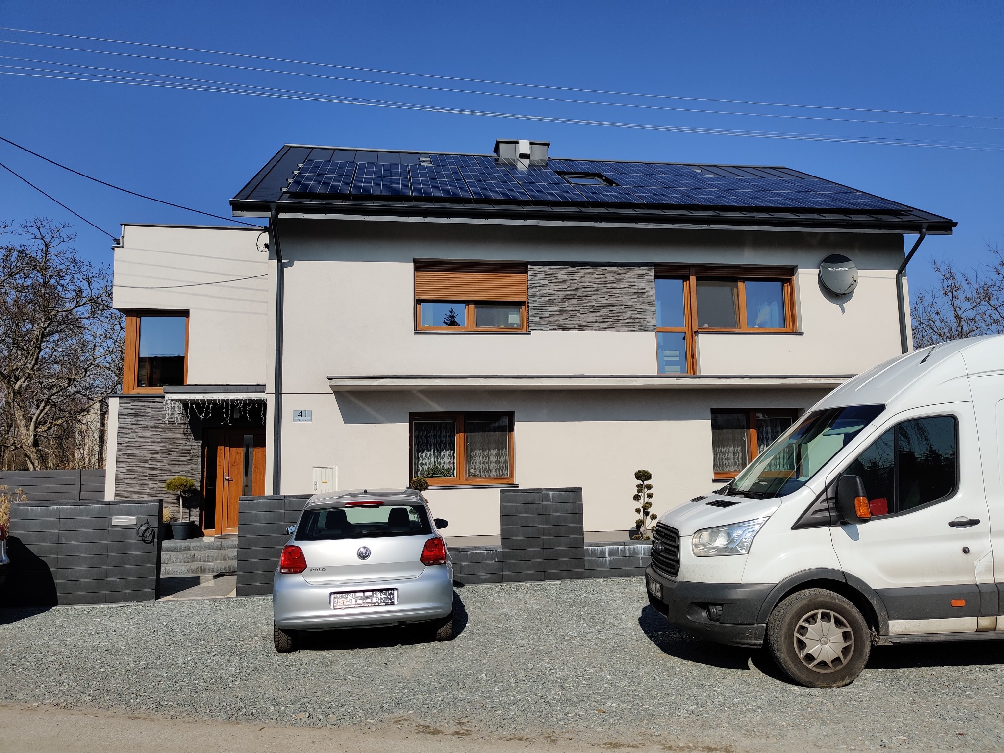 Dwupiętrowy dom z panelami słonecznymi na dachu, zaparkowany srebrny Volkswagen Polo i biały Ford Transit na żwirowym podjeździe w słoneczny dzień.