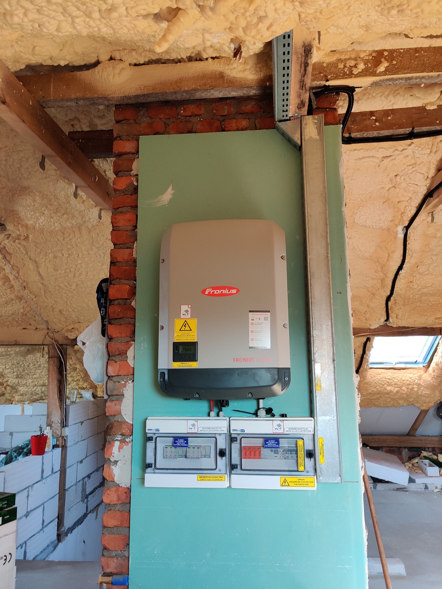 Szary falownik Fronius zamontowany na zielonej płycie, pod nim dwie skrzynki rozdzielcze, widoczne okablowanie, ściana z cegły i ocieplenie pianką na poddaszu.