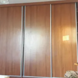 Duża szafa przesuwna z trzema drzwiami, z których jedno jest lustrzane, odbijające fragment pokoju z komodą i obrazami w ozdobnych ramach.