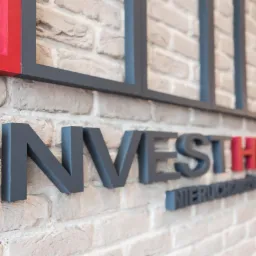 Szare, przestrzenne litery 'INVESTHOME' na ceglanej ścianie, z elementami graficznymi w kolorze czarnym i czerwonym. Ujęcie z boku, ukazujące fakturę cegieł.