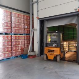 Luna24.pl - Pomarańczowy wózek widłowy STILL wjeżdża do magazynu z wysokimi regałami paletowymi wypełnionymi napojami w foliowych opakowaniach.