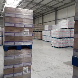 Wnętrze magazynu z paletami i kartonami owiniętymi folią, widoczne ślady kół na betonowej posadzce, metalowa konstrukcja dachu.