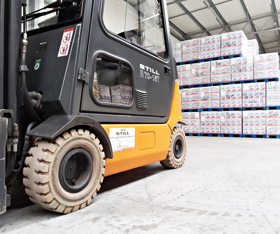 Wózek widłowy STILL R70-16T w magazynie z paletami zapakowanymi w kartony, widoczne opony wózka i część kabiny, w tle regały z towarem.