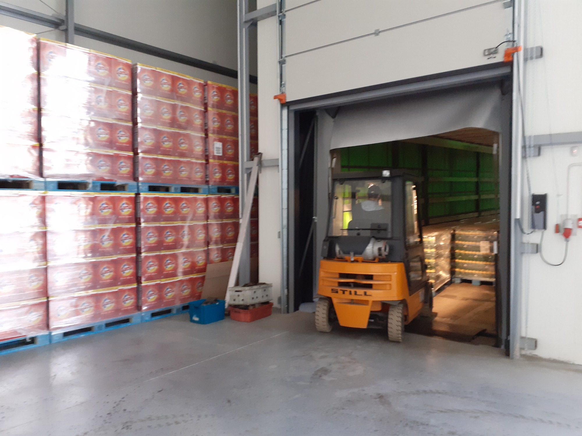 Pomarańczowy wózek widłowy STILL wjeżdża do magazynu z wysokimi regałami paletowymi wypełnionymi napojami w foliowych opakowaniach.