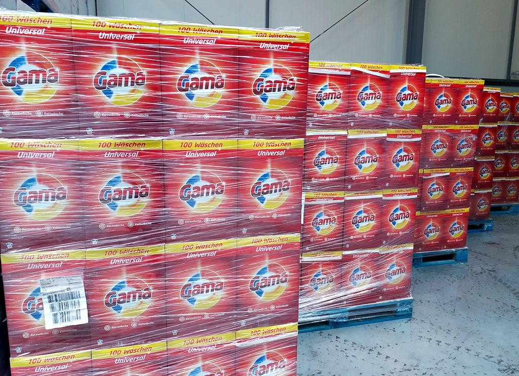 Palety zafoliowane stertami opakowań proszku do prania marki Gama Universal, ułożone w magazynie.