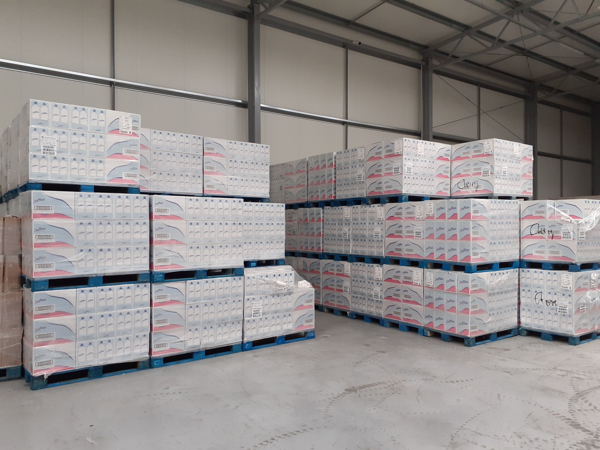 Palety z zapakowanymi kartonami płynu do płukania Softlan w magazynie z metalową konstrukcją, widoczne ślady opon na podłodze.
