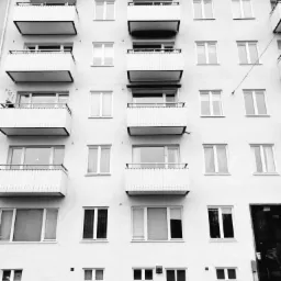 Fasada budynku mieszkalnego z rzędem balkonów, sfotografowana w czerni i bieli, z widocznym fragmentem chodnika przed budynkiem.