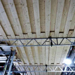 Sufit w trakcie montażu z widocznymi drewnianymi deskami i metalową konstrukcją podtrzymującą, widok z dołu z rusztowaniami w tle.