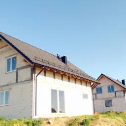 Dwa surowe domy jednorodzinne z szarym dachem i czarnymi rynnami, widoczne bloczki konstrukcyjne i drewniane elementy więźby dachowej, w tle błękitne niebo.