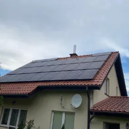 Panele słoneczne na dachu domu jednorodzinnego w Olsztynie, widok z dołu na instalację fotowoltaiczną, z kominem w tle i fragmentem błękitnego nieba z chmurami.