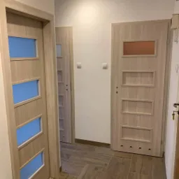 Korytarz z trzema zamontowanymi drzwiami wewnętrznymi w odcieniach drewna, z panelami szklanymi w dwóch parach drzwi i pełną płytą w trzeciej parze, na podłodze brązowe płytki imitujące drewno...