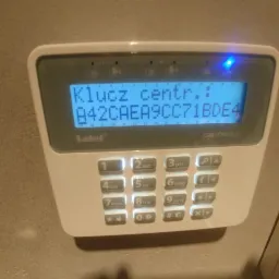 Biały panel sterowania alarmu firmy Satel, model Perfecta, z wyświetlaczem pokazującym klucz centrali i niebieską diodą LED, zamontowany na ścianie.