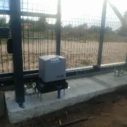 Szary automat FAAC 740 zamontowany na betonowej podstawie przy przesuwnej bramie z metalowej siatki, widoczne śruby mocujące i elementy instalacji.