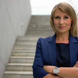 Portret kobiety w granatowym żakiecie, skrzyżowane ręce, zegarek na nadgarstku, na tle schodów i szklanej balustrady. Profesjonalny wizerunek.