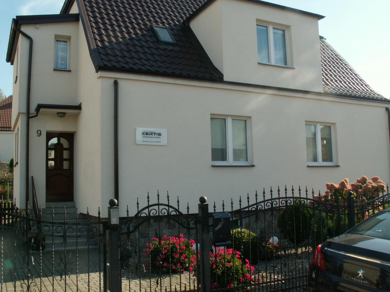 Dwupiętrowy budynek z brązowym dachem i beżową elewacją, numer 9 przy wejściu, tabliczka z napisem CERTIS, ozdobne metalowe ogrodzenie z czerwonymi kwiatami i fragment zaparkowanego samochodu.