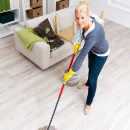 Kobieta w żółtych rękawicach czyści podłogę mopem w jasnym salonie z beżową sofą i białymi regałami, patrząc w obiektyw.
