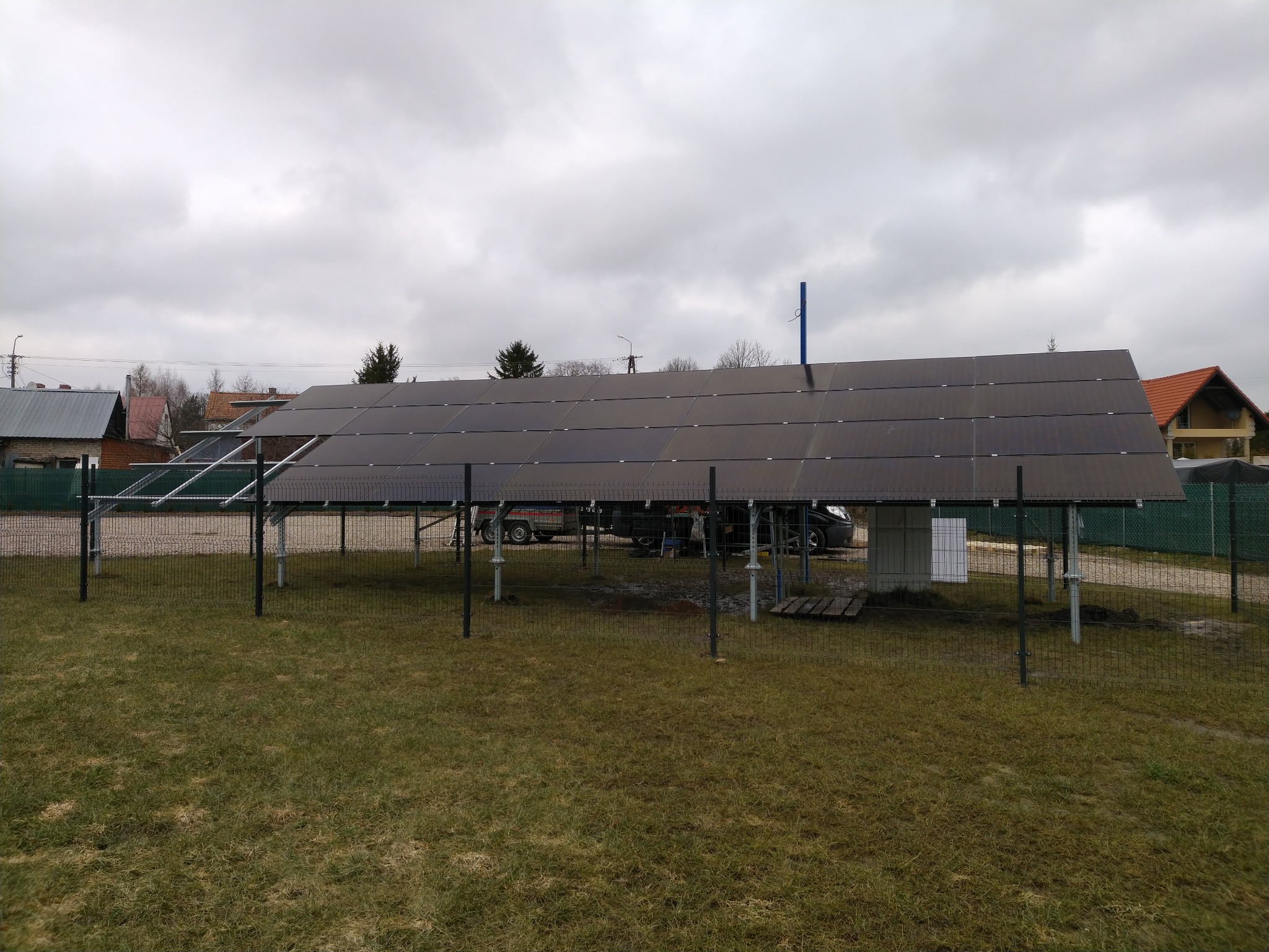 Panele słoneczne zamontowane na metalowej konstrukcji na trawiastym terenie, widoczne zabudowania w tle, pochmurne niebo.