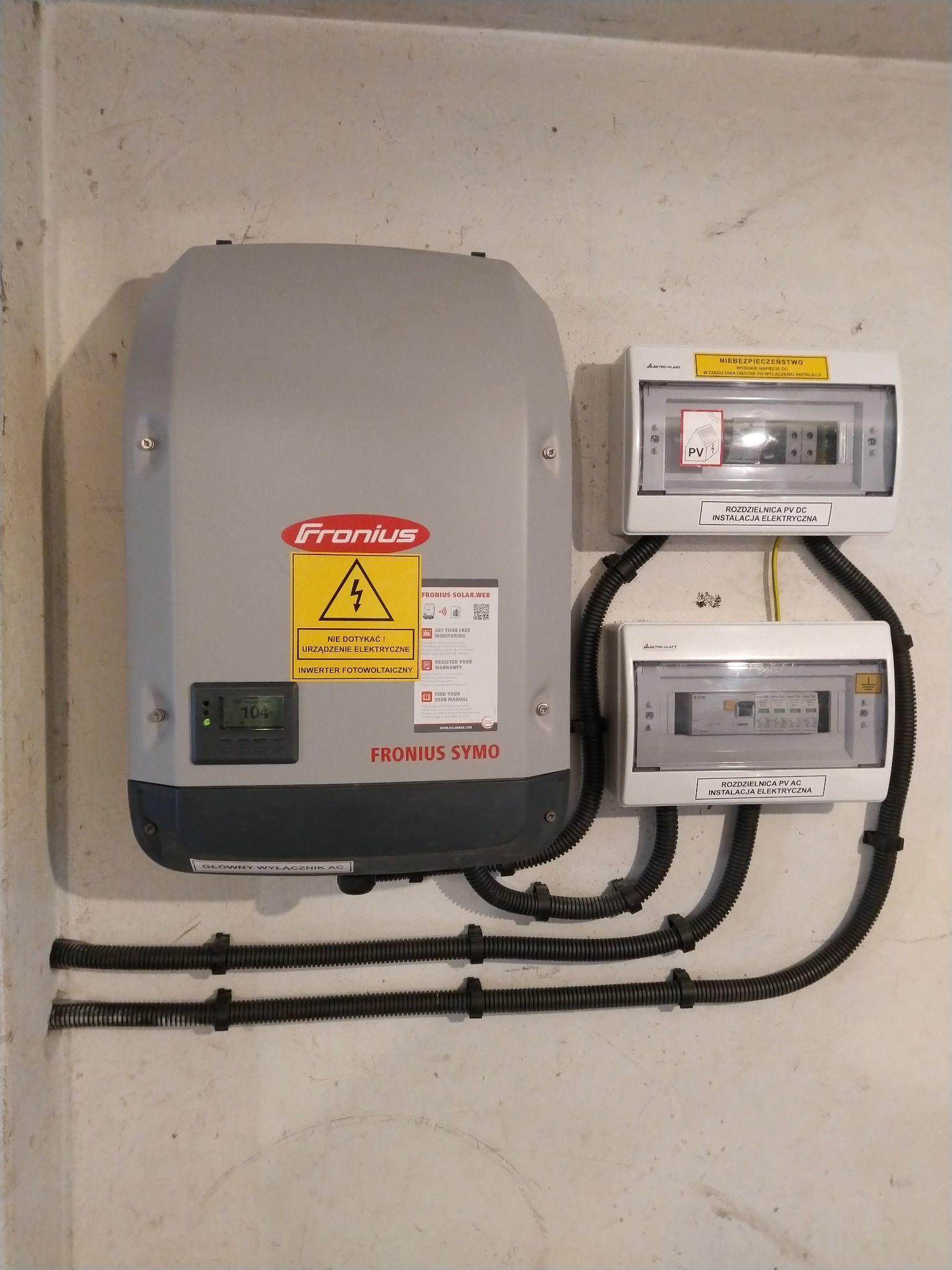 Szary falownik Fronius zamontowany na ścianie z dwoma skrzynkami rozdzielczymi PV AC i PV DC, połączony rurami karbowanymi. Widoczny wyświetlacz falownika z wartością 104.