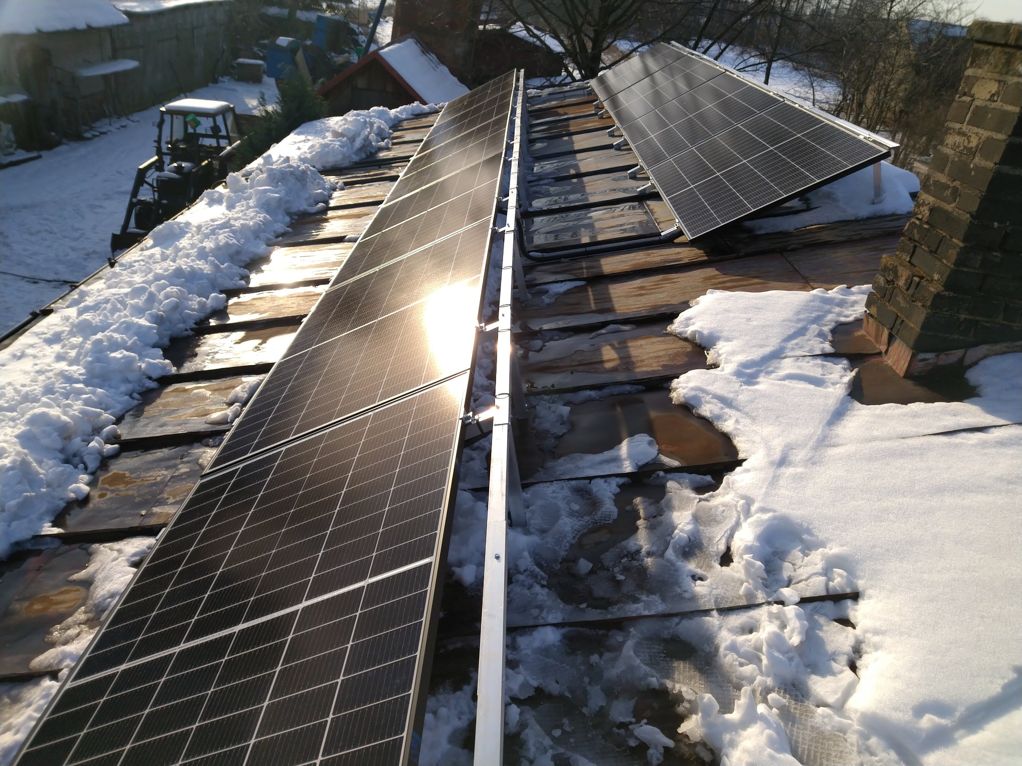 Panele fotowoltaiczne na zaśnieżonym dachu z ceglanym kominem w słoneczny dzień, w tle widoczny traktor odśnieżający.