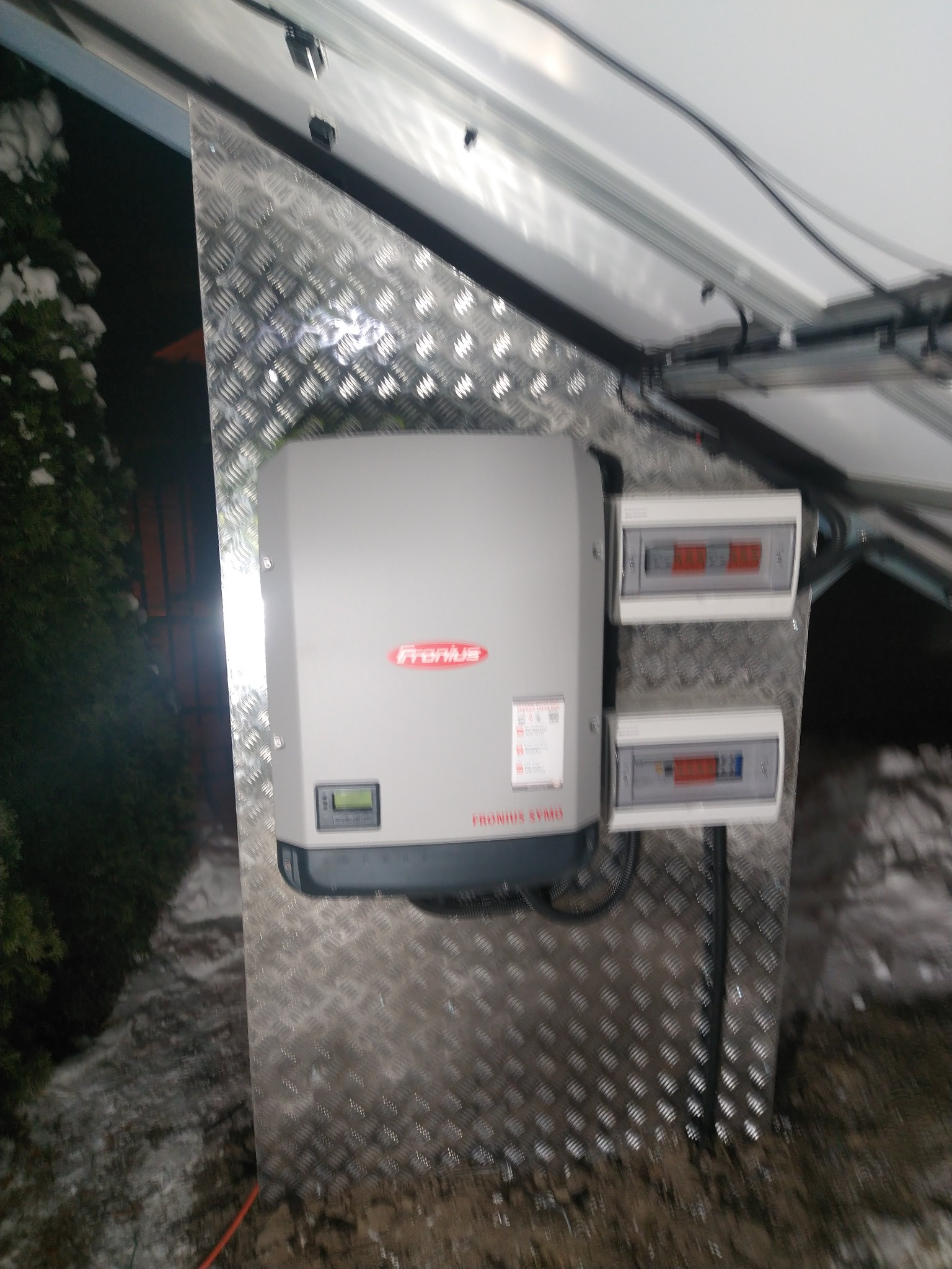 Szary inwerter Fronius Symo zamontowany na aluminiowej płycie ryflowanej, z widocznymi dwoma skrzynkami zabezpieczeń, instalacja w warunkach zimowych, śnieg na roślinności w tle.