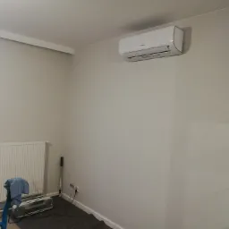 Biała jednostka klimatyzacji zamontowana na ścianie w jasnym pokoju, widoczny grzejnik, okno i narzędzia sugerujące niedawny montaż.