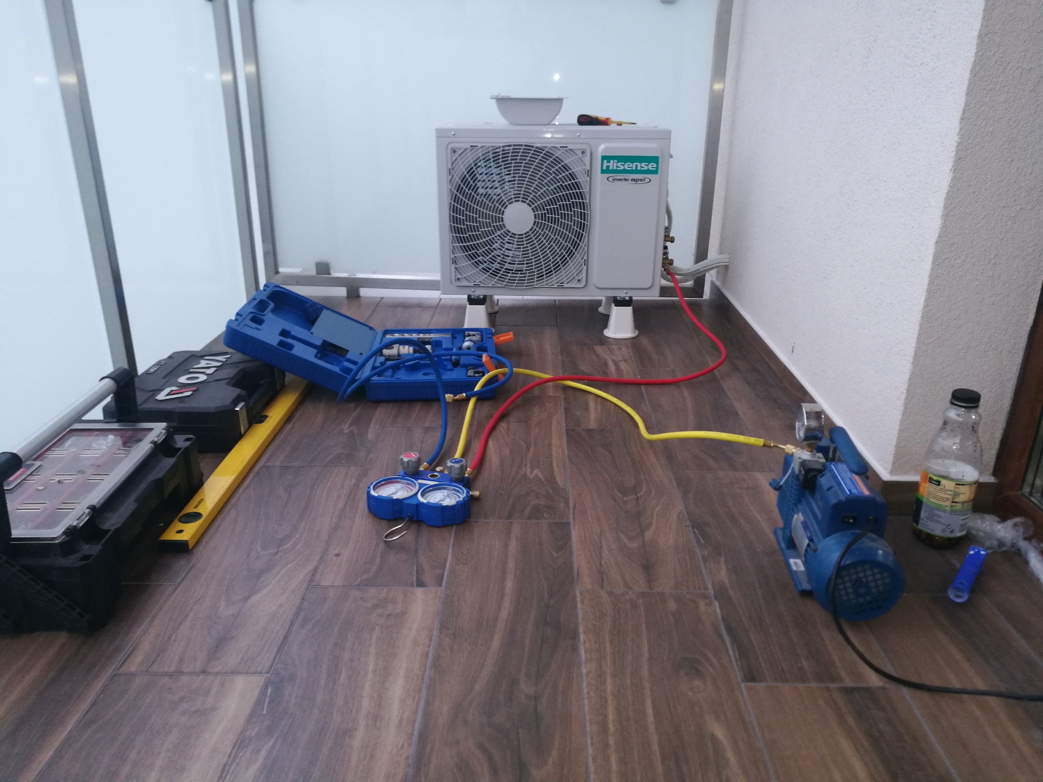 Biała jednostka zewnętrzna klimatyzacji Hisense, ustawiona na balkonie z drewnianą podłogą, podłączona do niebieskiej pompy próżniowej i manometrów za pomocą żółtych i czerwonych węży, obok...