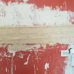 Fragment czerwonej ściany w trakcie dekoracyjnego malowania z efektem cegieł, widoczne ślady wcześniejszych warstw farby i gniazdko elektryczne z wystającymi kablami.