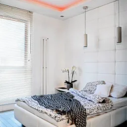 Nowoczesna sypialnia z białym łóżkiem, pikowanym zagłówkiem, podwieszanymi lampami i subtelnym oświetleniem LED na suficie; czarne szpilki leżące na podłodze dodają elegancji.