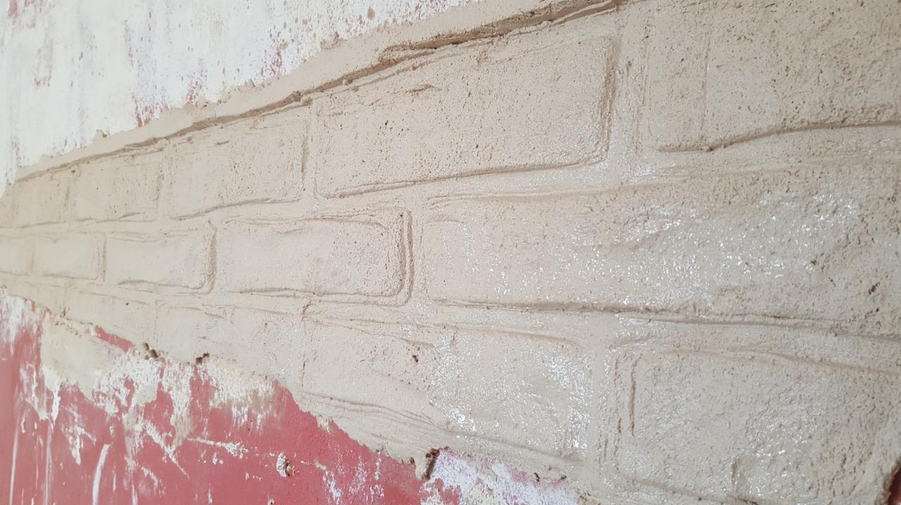 Ściana z imitacją cegieł, świeżo pokryta beżową zaprawą, widoczne niedociągnięcia przy czerwonej farbie dolnej części ściany.