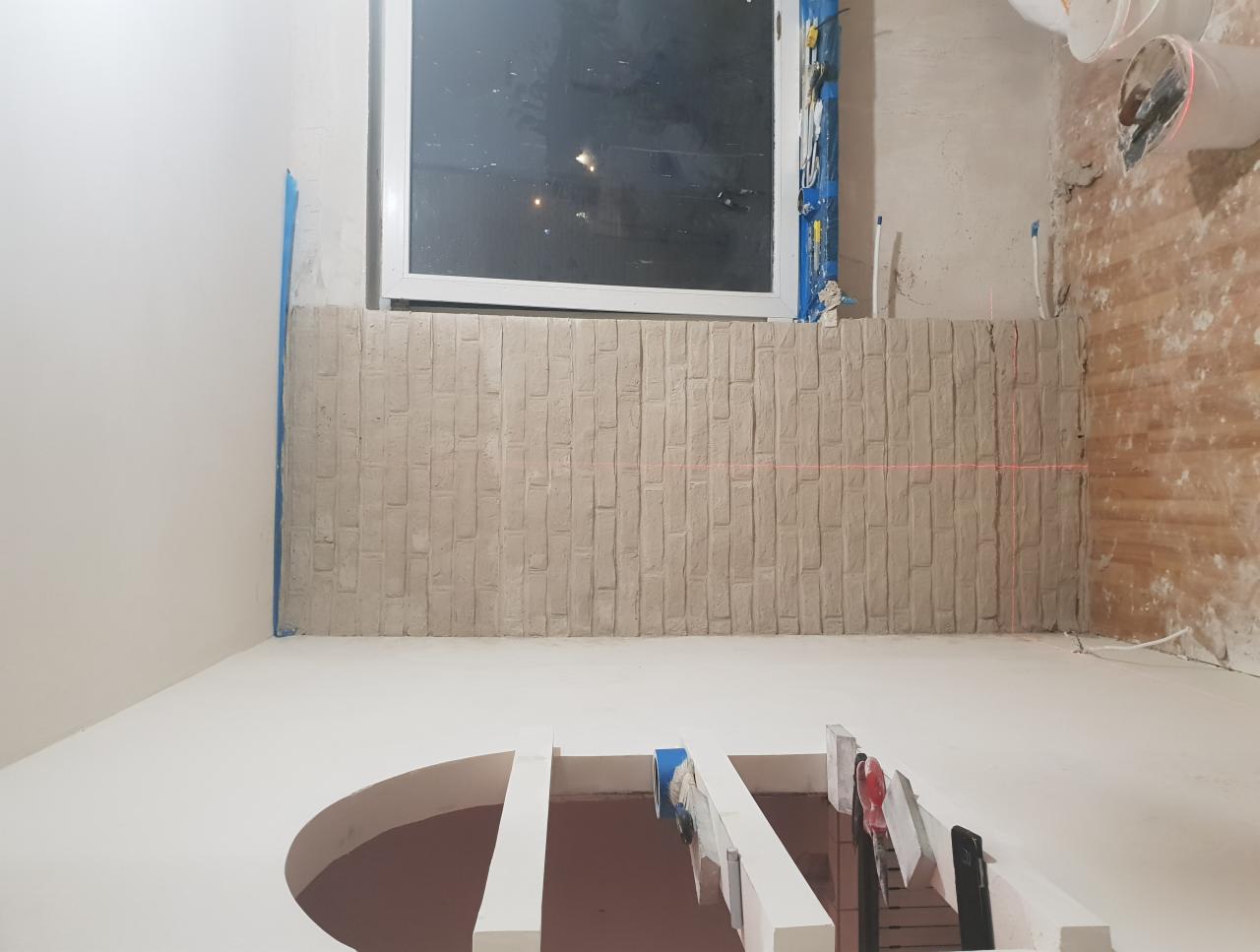 Ściana w trakcie wykończenia imitująca cegłę, z widoczną poziomą linią lasera, fragment podłogi z elementami konstrukcyjnymi w pomieszczeniu z oknem.