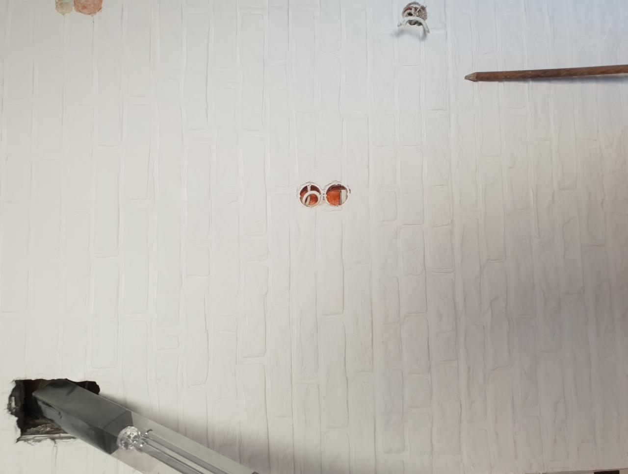 Ściana z fakturą cegieł pomalowana na biało, widoczne puszki elektryczne oraz otwór z wystającym kablem i fragment drabiny.