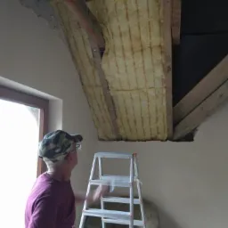 Mężczyzna w czapce na drabinie, spogląda na izolację z wełny mineralnej w skośnym suficie poddasza, widoczne drewniane belki konstrukcyjne i fragment okna.