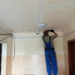 Instalator w niebieskim kombinezonie na drabinie, montuje kamerę monitoringu pod sufitem korytarza w budynku mieszkalnym, widoczne kable i listwy montażowe.