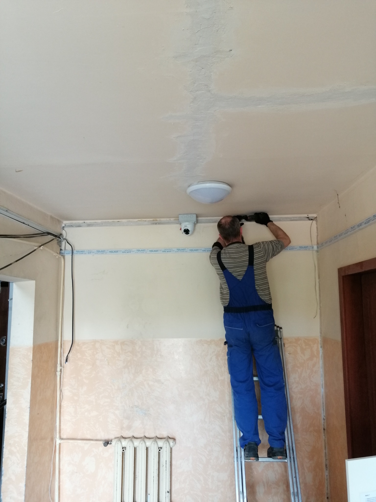 Instalator w niebieskim kombinezonie na drabinie, montuje kamerę monitoringu pod sufitem korytarza w budynku mieszkalnym, widoczne kable i listwy montażowe.