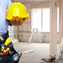 Pracownik budowlany trzyma żółty kask i młotek, w tle surowy stan pomieszczenia z drewnianą konstrukcją ścian i dwiema drabinkami.