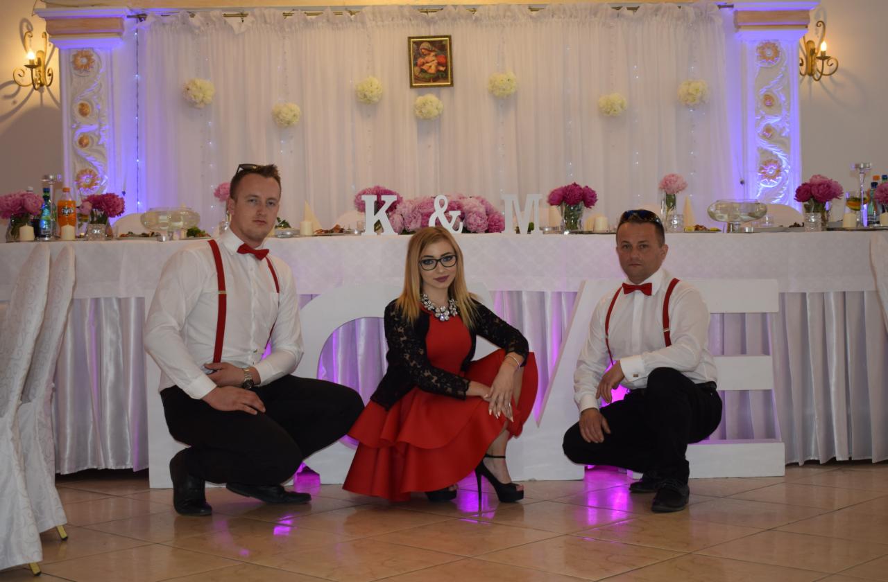Dwaj mężczyźni w białych koszulach z czerwonymi szelkami i muchami oraz kobieta w czerwonej sukience i czarnym żakiecie pozują na tle dekoracji ślubnej z białymi kwiatami i literami K&M.