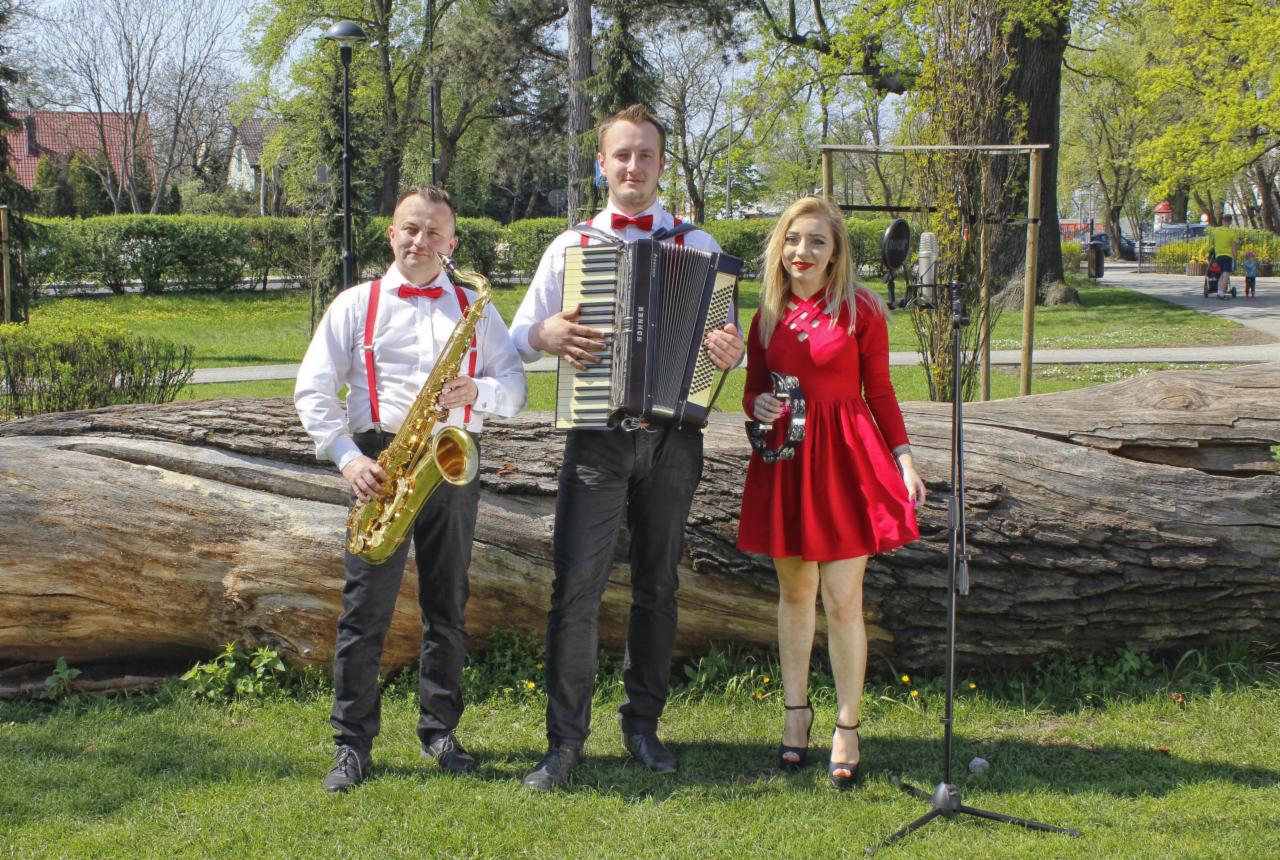 Trzyosobowy zespół w plenerze: saksofonista z czerwonymi szelkami i muszką, akordeonista w białej koszuli z muszką i wokalistka w czerwonej sukience z tamburynem, pozują na tle powalonego pnia...