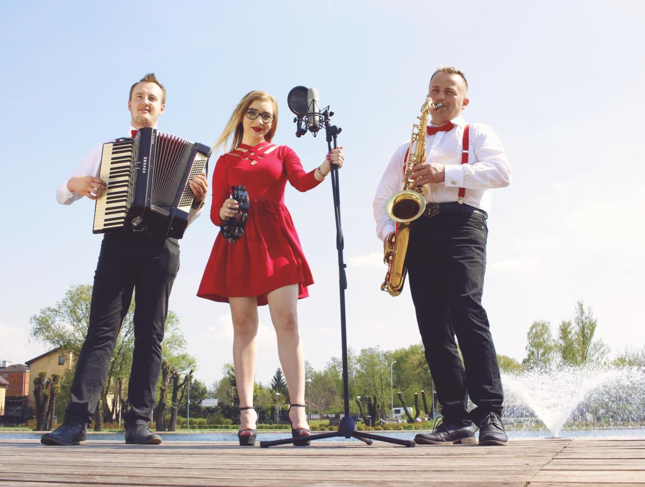 Trzyosobowy zespół muzyczny na drewnianym pomoście nad jeziorem, wokalistka w czerwonej sukience z tamburynem, akordeonista i saksofonista w koszulach z muszkami i szelkami, w tle fontanna.