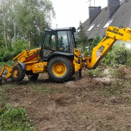 Żółta koparko-ładowarka JCB na placu budowy, widoczne ślady pracy w postaci wykopanej ziemi i resztek roślinności, w tle dom z szarą dachówką i zieleń drzew.