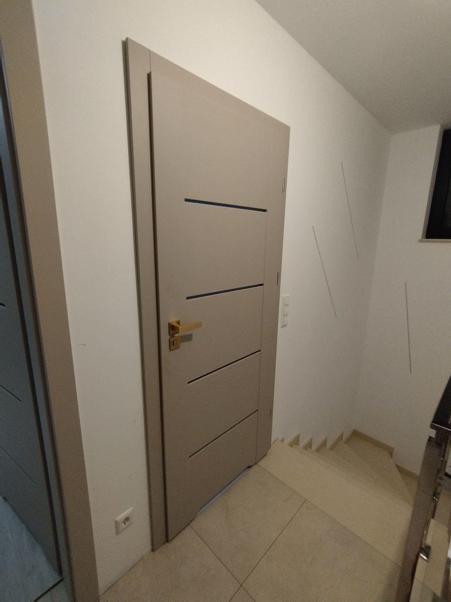 Beżowe drzwi wewnętrzne z poziomymi frezami i złotą klamką, zamontowane w jasnym wnętrzu z widocznymi schodami i fragmentem innej zabudowy.