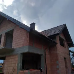 Dom w trakcie budowy z częściowo ukończonym pokryciem dachowym z dachówki ceramicznej i zamontowanymi rynnami w kolorze grafitowym, widoczne rusztowanie i mur z czerwonej cegły.