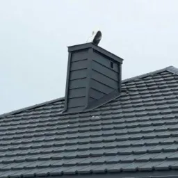 Szary dach z dachówki ceramicznej z blachy trapezowej, komin obłożony blachą, w tle pochmurne niebo.