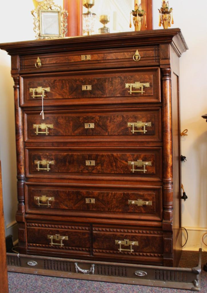 Antyczna, drewniana komoda z sześcioma szufladami, ozdobiona mosiężnymi uchwytami i detalami, stojąca na tle ściany i dekoracyjnego grzejnika.