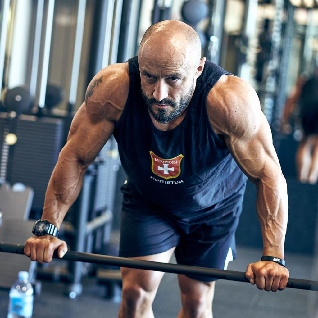 Mężczyzna z brodą, w czarnej koszulce z logo i tatuażem na ramieniu, ćwiczy z obciążeniem na siłowni, widoczne napięte mięśnie i butelka wody.